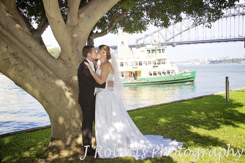 Bride and groom kissing under a tree with Sydney Ferry in the background - wedding photography sydney